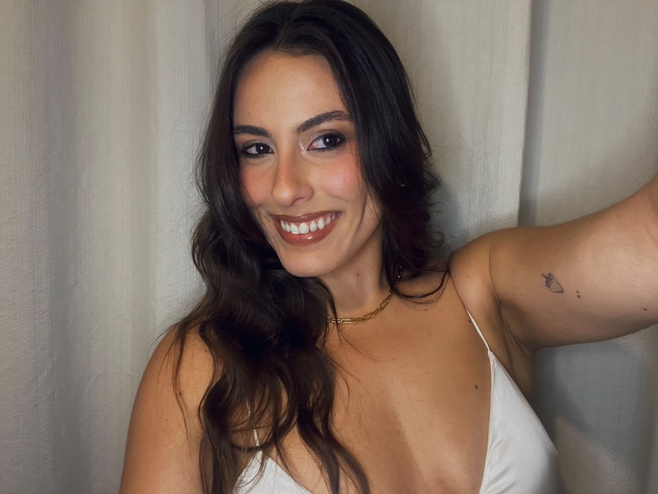 Bettina Poll sorrindo em selfie com blusa de cetim clara e colar dourado